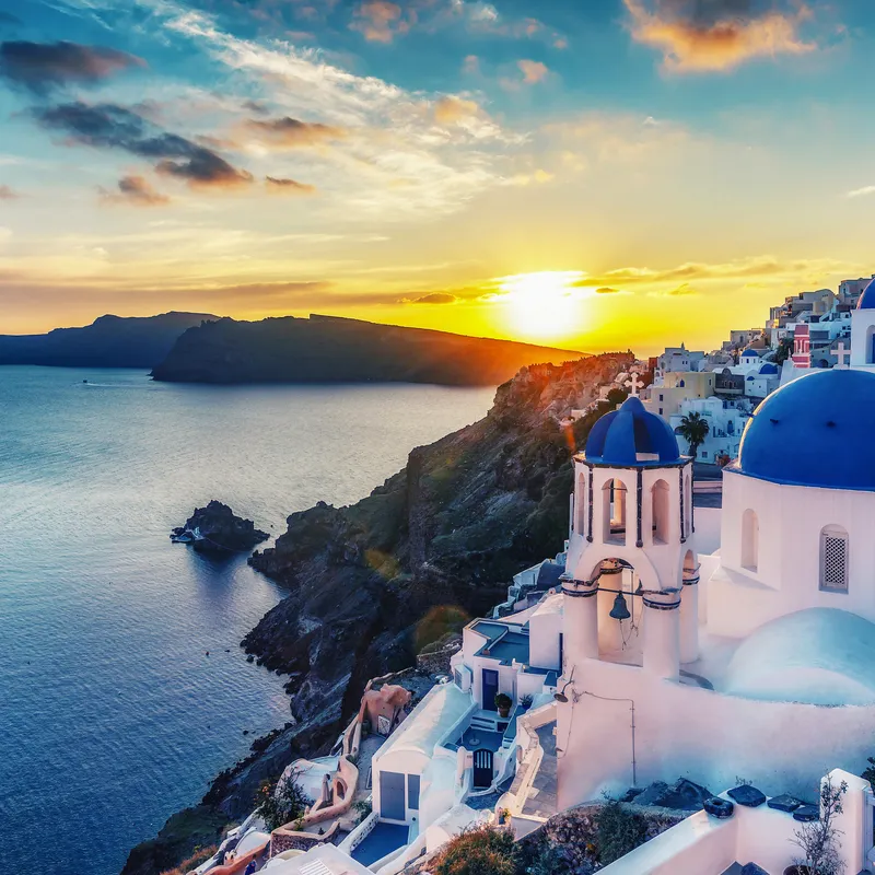 Panoramablick auf Oia, Santorin bei Sonnenuntergang mit blau-weißer Kirche im Vordergrund.