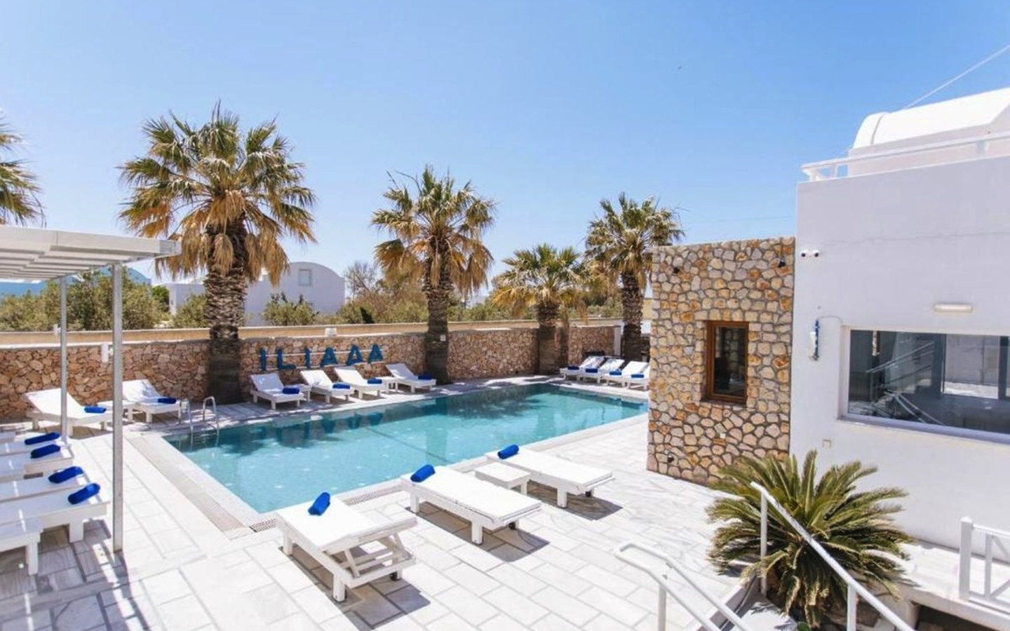 Poolbereich des Iliada Odysseas Resorts in Santorin, umgeben von Palmen und Liegestühlen.