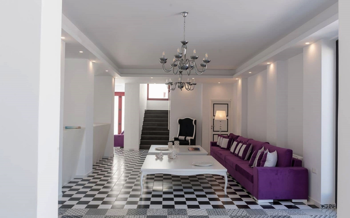 Helle Lounge mit lila Sofa, weißen Wänden und Schachbrettboden, Kronleuchter oben.