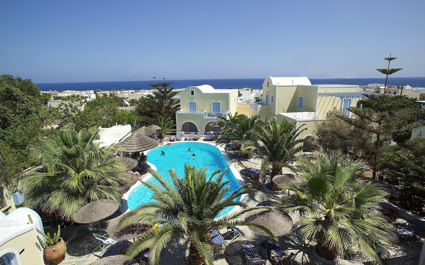 Hotelpool umgeben von Palmen mit Blick auf weißen Gebäuden und das Meer in Santorin.