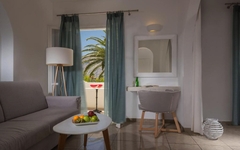 Wohnbereich eines hellen Hotelzimmers mit Sofa, Tisch und Blick auf Palmen.