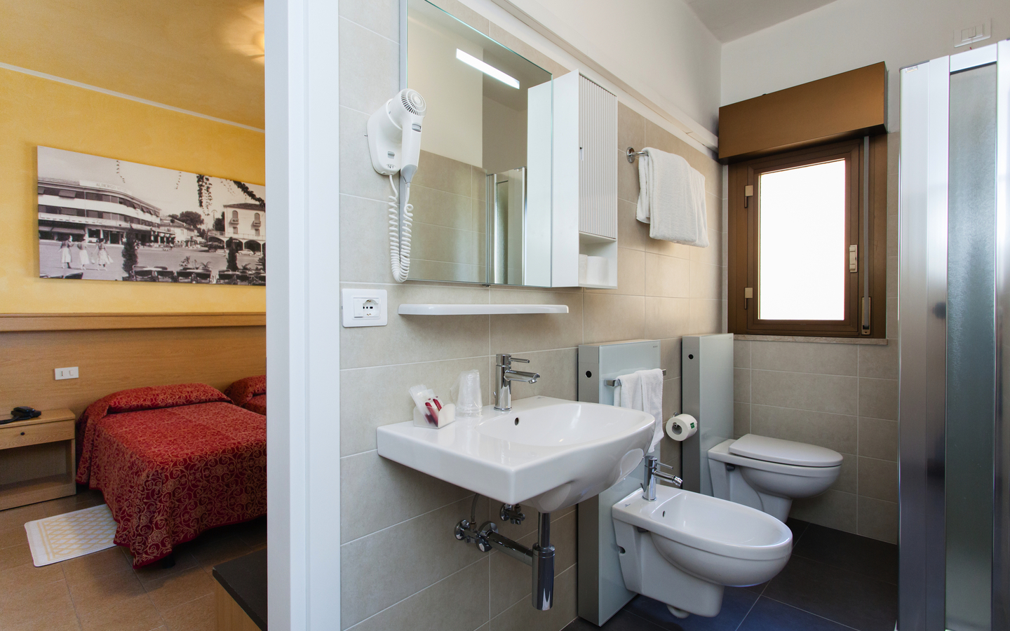 Badezimmer eines Hotelzimmers mit Waschbecken, Bidet und Dusche, im Hintergrund ein Bett.