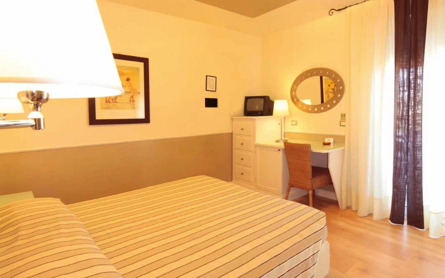 Hotelzimmer mit Doppelbett, Schreibtisch, Spiegel und Lampe in warmem Licht.