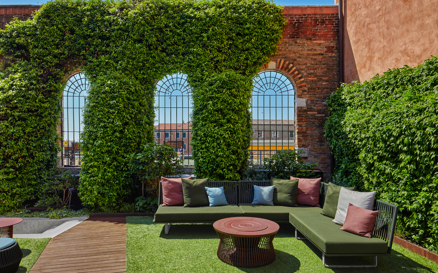 Lounge-Bereich im Freien mit Sitzsofas und Kissen vor Ziegelmauer und Pflanzen.