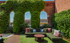 Lounge-Bereich im Freien mit Sitzsofas und Kissen vor Ziegelmauer und Pflanzen.
