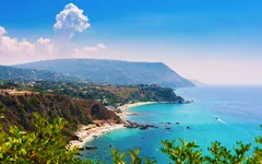 Küste mit klarem, türkisfarbenem Wasser und bewaldeten Klippen unter blauem Himmel.