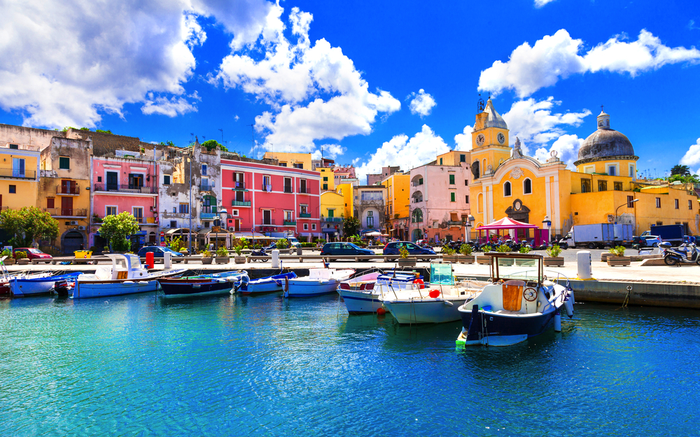 Bunte Häuser und Boote am Hafen von Procida, unter blauem Himmel.