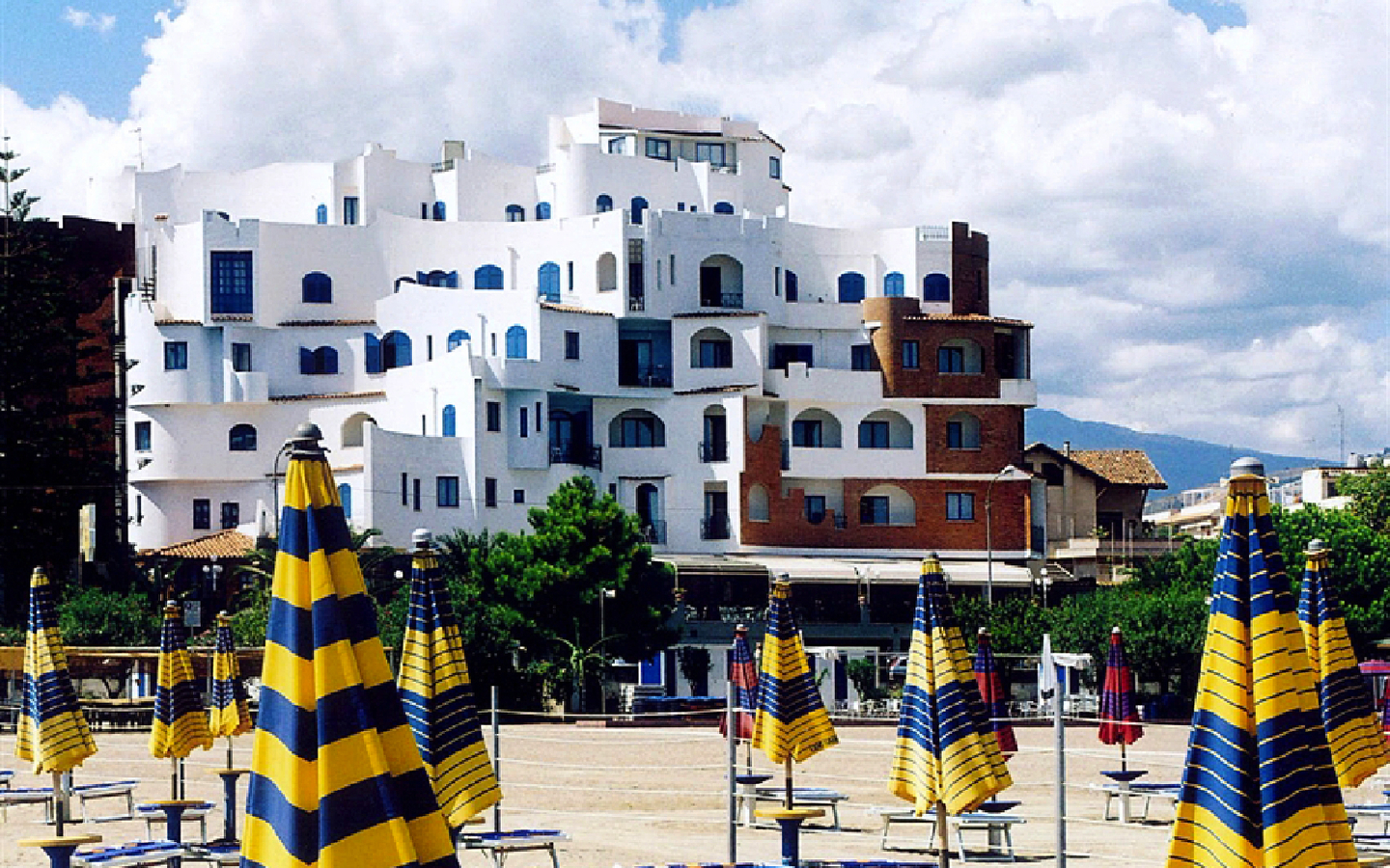 Hotel am Strand mit gestreiften Sonnenschirmen, dahinter Gebäude und Bäume.