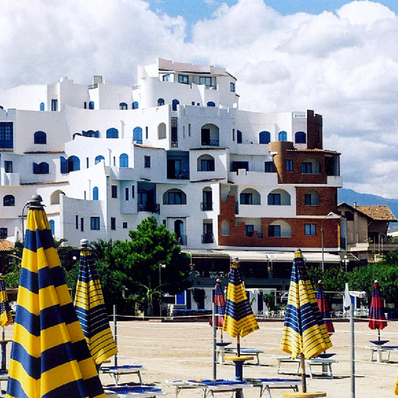 Hotel am Strand mit gestreiften Sonnenschirmen, dahinter Gebäude und Bäume.