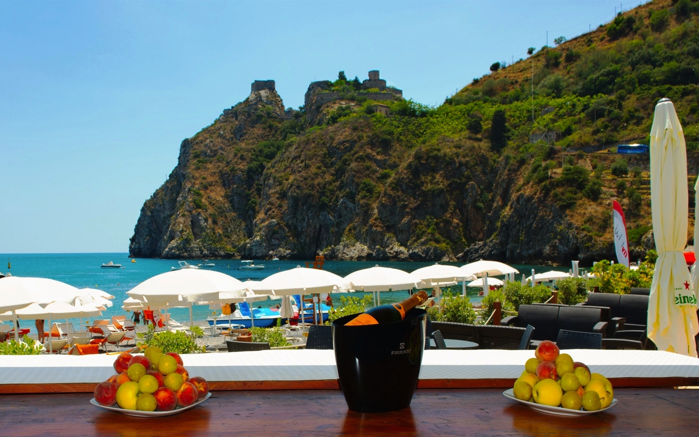 Blick von einer Terrasse auf einen Strand mit Felsen und Sonnenschirmen in Sizilien.