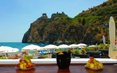 Blick von einer Terrasse auf einen Strand mit Felsen und Sonnenschirmen in Sizilien.