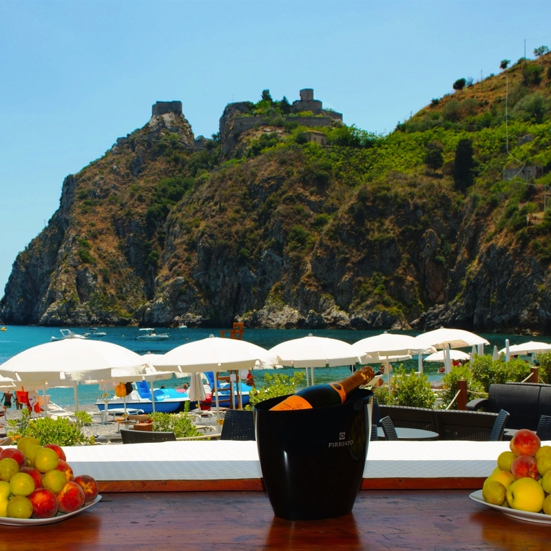 Blick von einer Terrasse auf einen Strand mit Felsen und Sonnenschirmen in Sizilien.