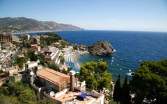 Blick auf Küste und Gebäude von Taormina, Sizilien, mit Meer und Hügeln im Hintergrund.