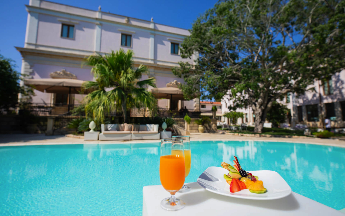 Hotelpool mit Palmen, orangefarbenes Getränk und Obstteller im Vordergrund.