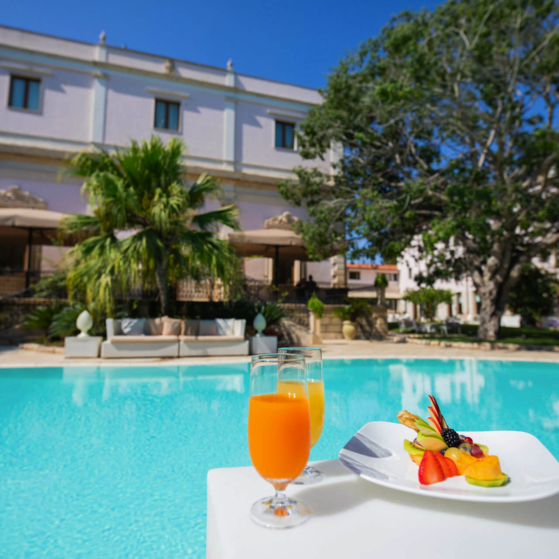 Hotelpool mit Palmen, orangefarbenes Getränk und Obstteller im Vordergrund.
