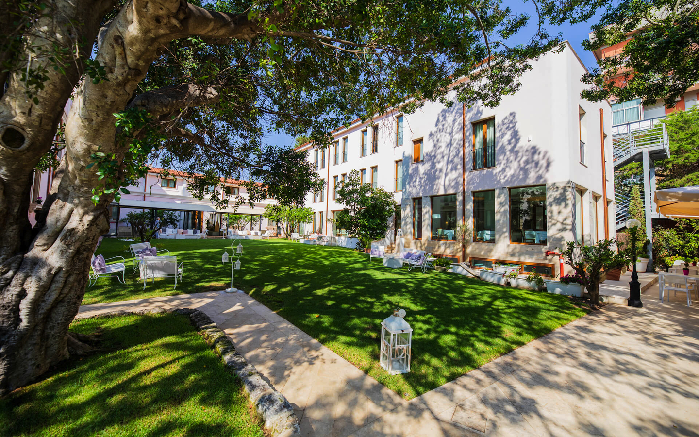 Außenansicht eines Hotels mit Garten und Bäumen im Sonnenschein.