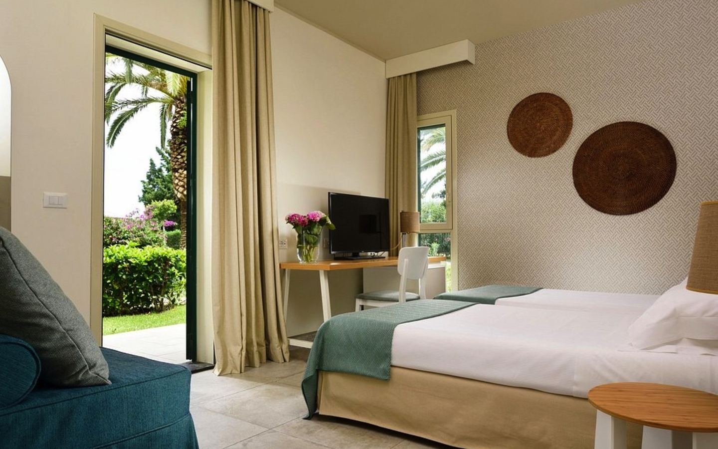 Hotelzimmer mit Doppelbett, Schreibtisch, Fernseher und Blick auf Garten mit Palmen.