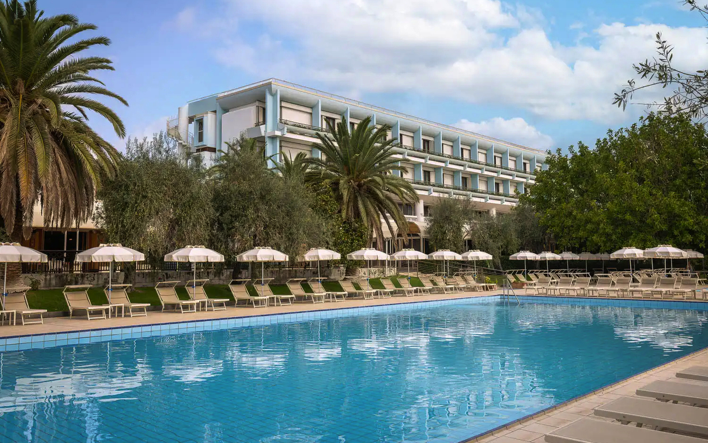 Außenansicht eines Hotels mit Palmen und großem Pool im Vordergrund.