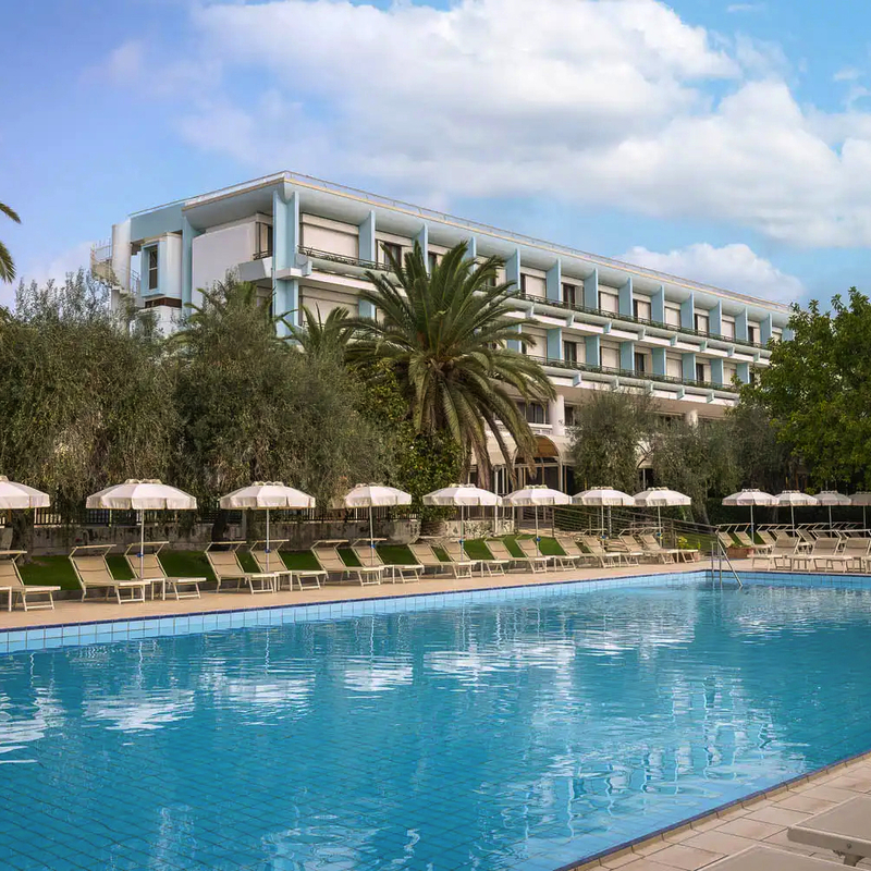 Außenansicht eines Hotels mit Palmen und großem Pool im Vordergrund.