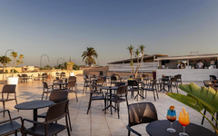 Offene Terrasse mit mehreren Tischen und Stühlen, dahinter eine Bar.