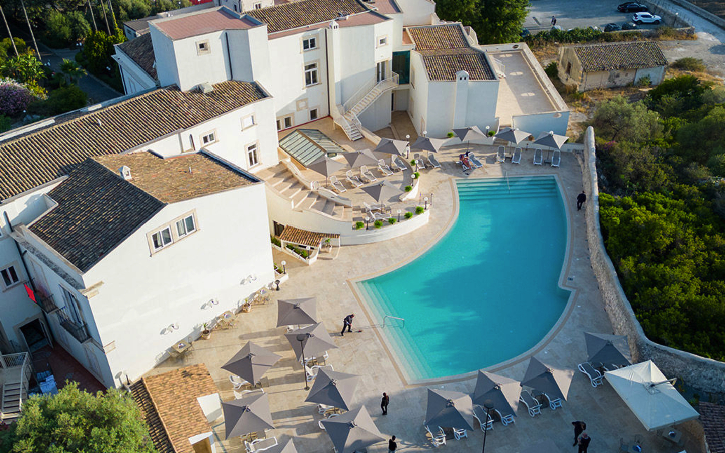 Hotelvilla mit großem Pool und Sonnenschirmen, umgeben von Bäumen und Parkplätzen.