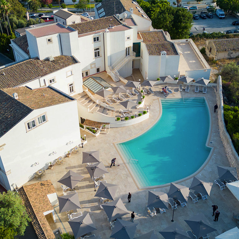 Hotelvilla mit großem Pool und Sonnenschirmen, umgeben von Bäumen und Parkplätzen.