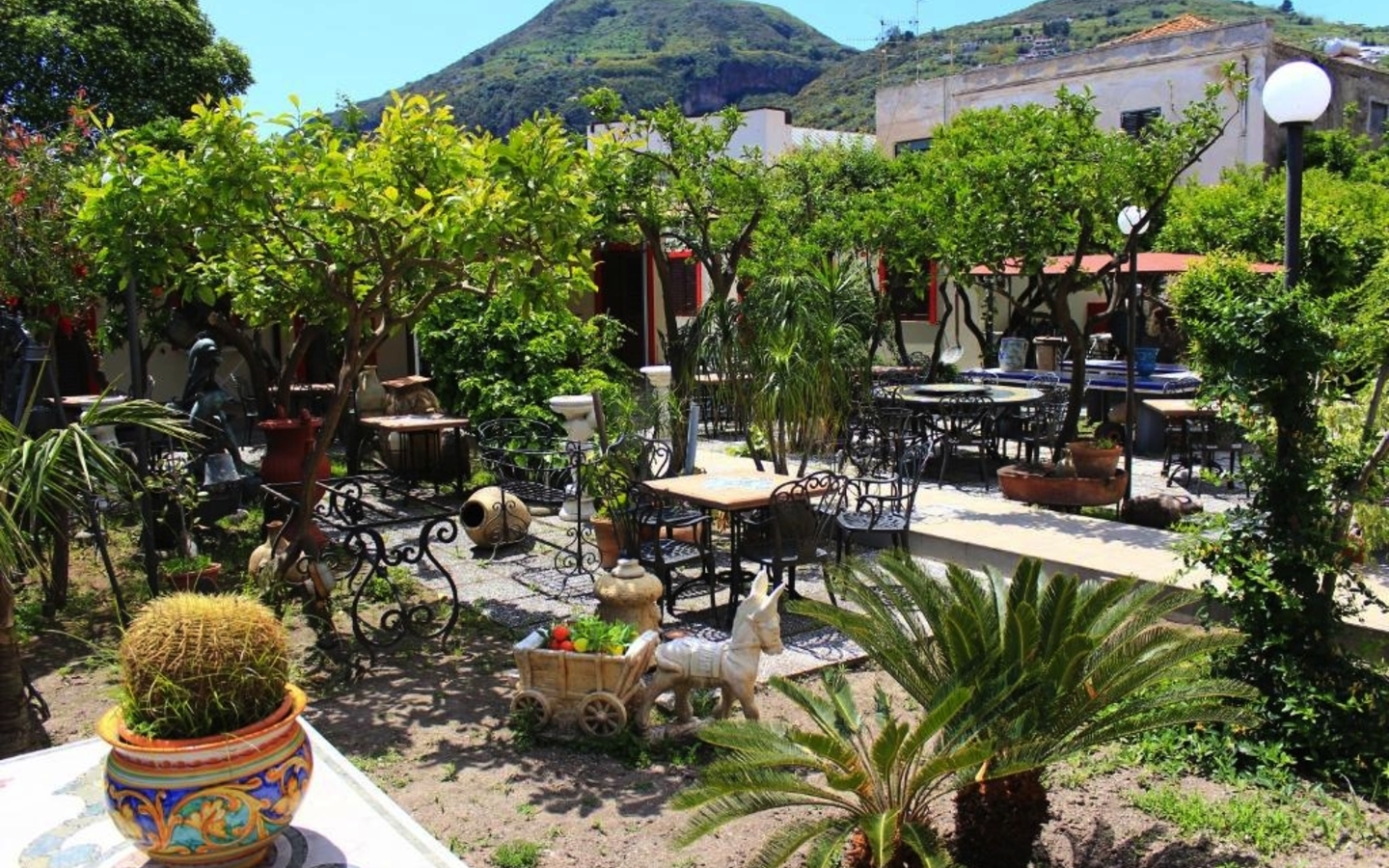 Garten mit Tischen und Stühlen, umgeben von Pflanzen und einem Hügel im Hintergrund.