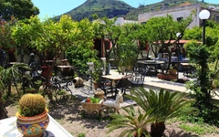 Garten mit Tischen und Stühlen, umgeben von Pflanzen und einem Hügel im Hintergrund.