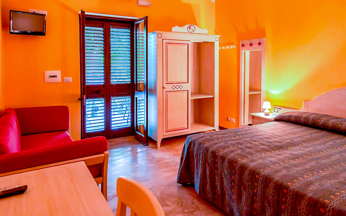 Hotelzimmer mit Doppelbett, Sofa, Kleiderschrank und Balkon in warmen Farben.