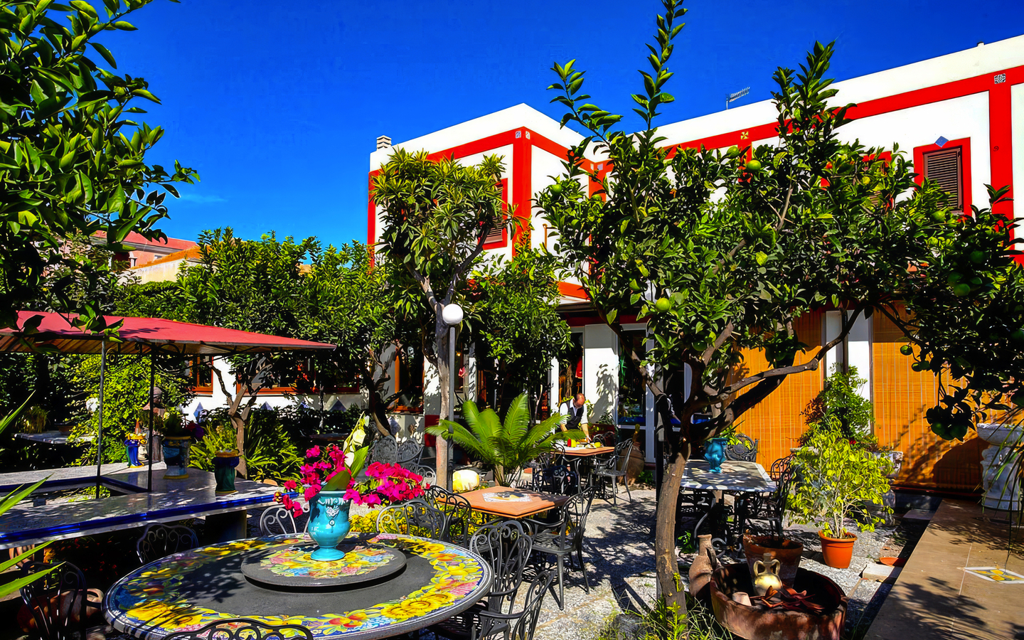 Garten eines Hotels mit bunten Tischen und üppiger Vegetation bei sonnigem Wetter
